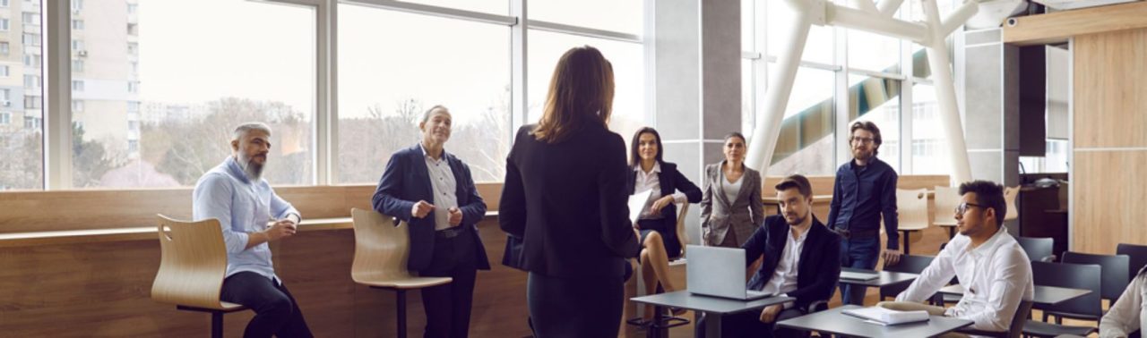 Various businessmen listen to female business leader during corporate meeting or training lecture.